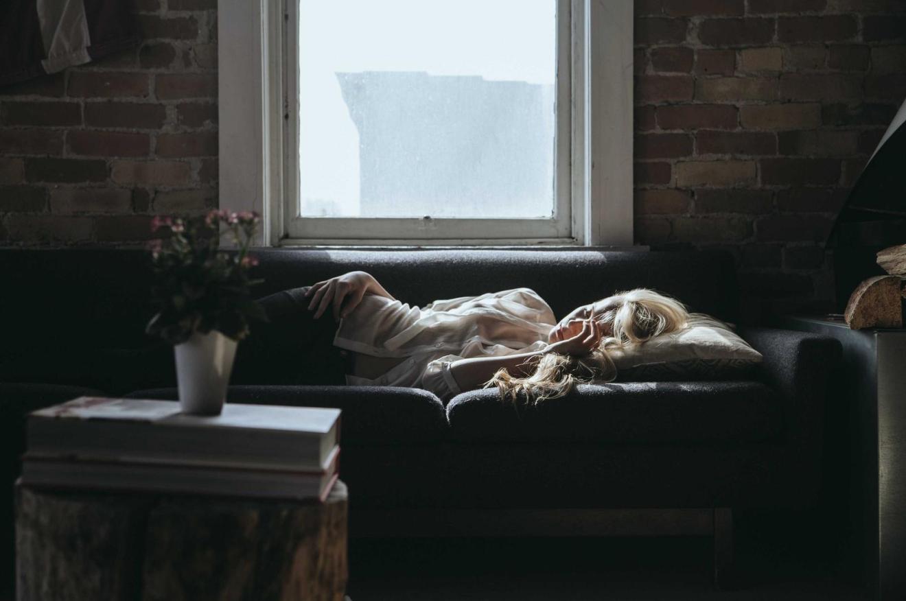 A person asleep on a sofa below a window in a dark brick room