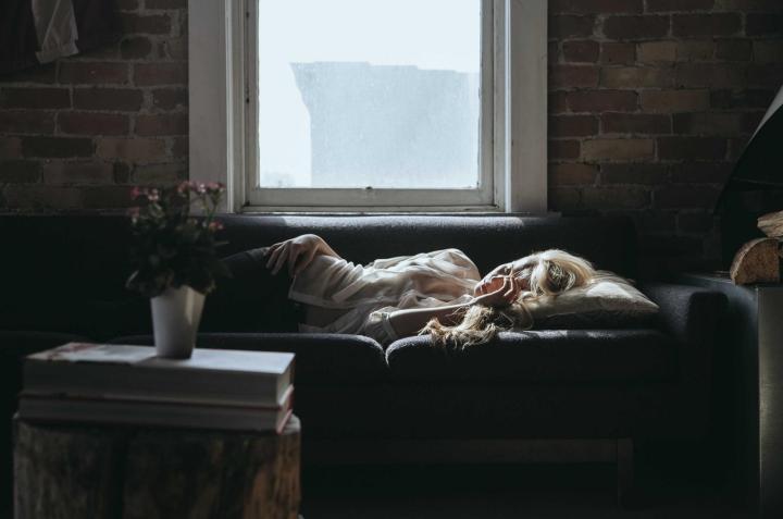 A person asleep on a sofa below a window in a dark brick room