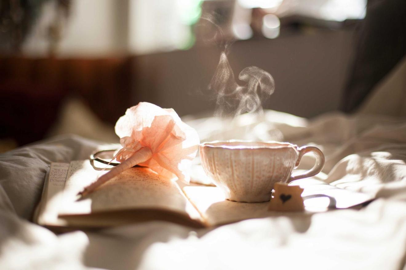 A steaming cup of tea beside an open book on a bed